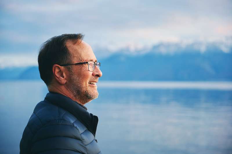 Older man standing outside with water and mountains in the background