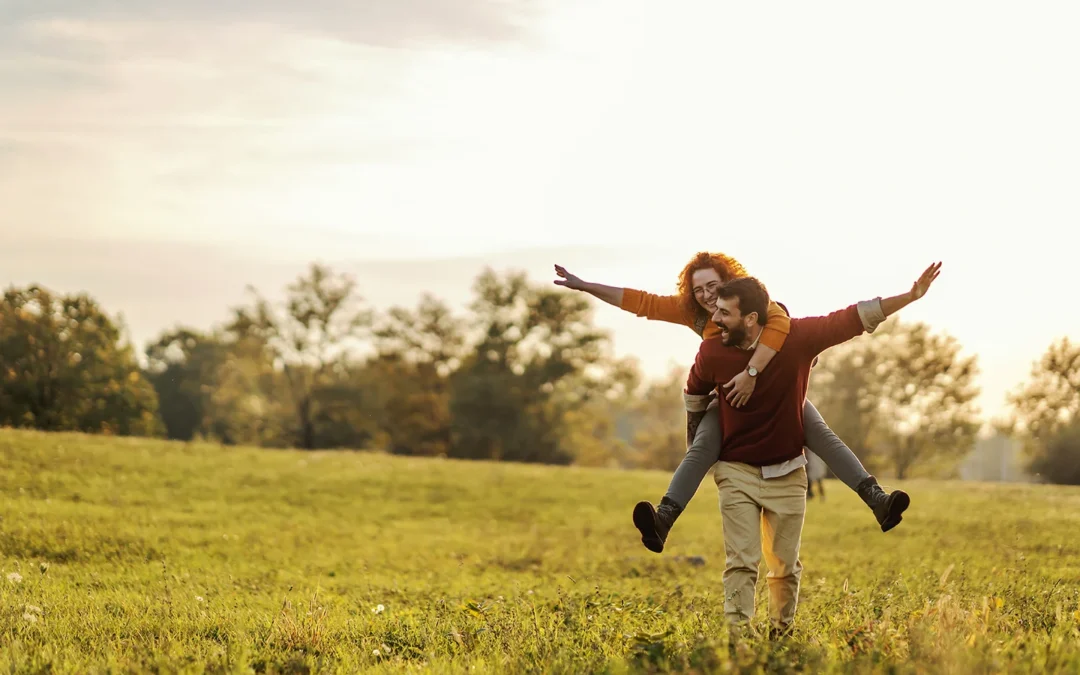 Happy young man giving a piggyback ride to a smiling woman in a sunlit grassy field.